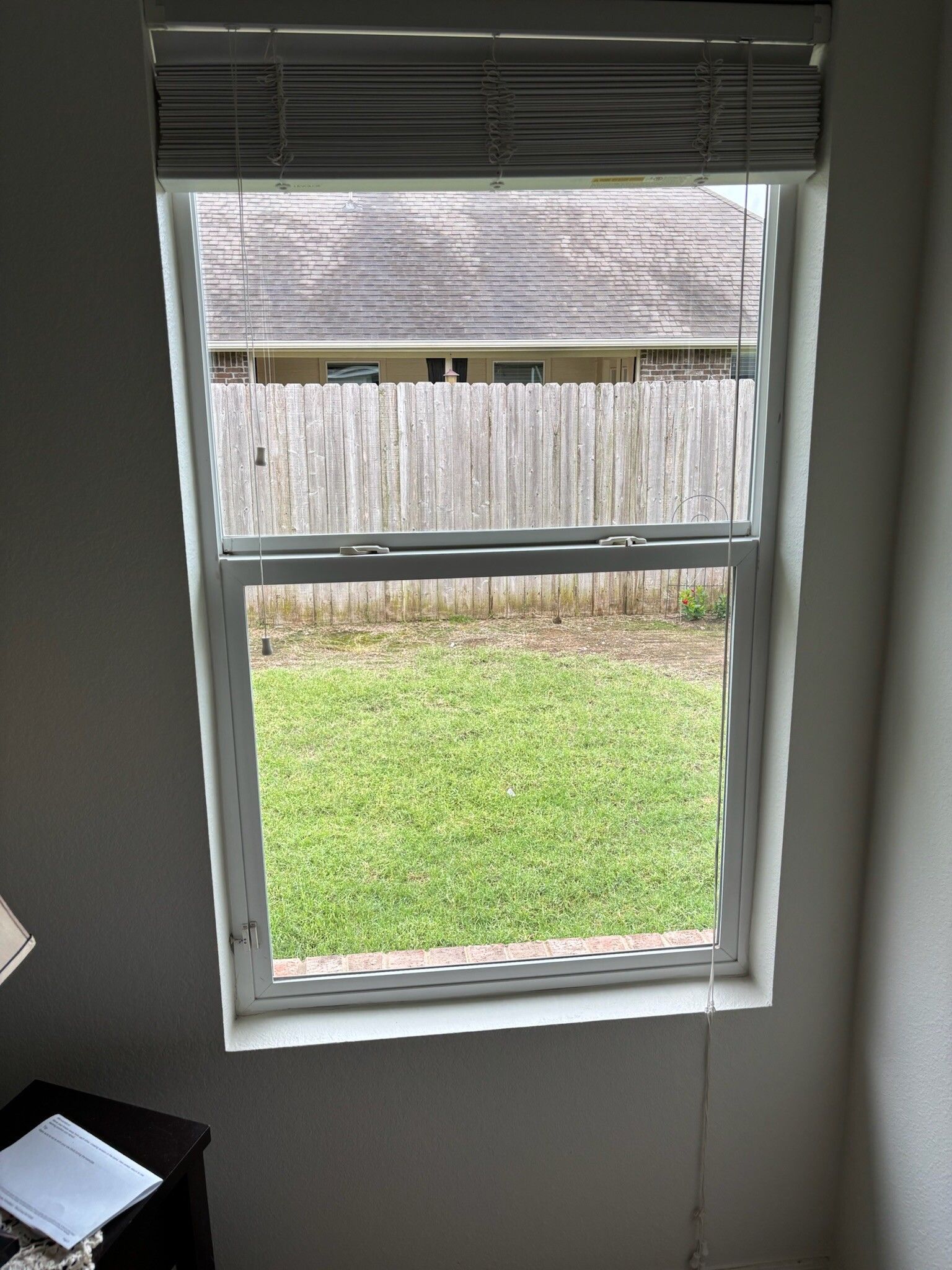 Window with blinds partially open, overlooking a grassy yard and wooden fence.
