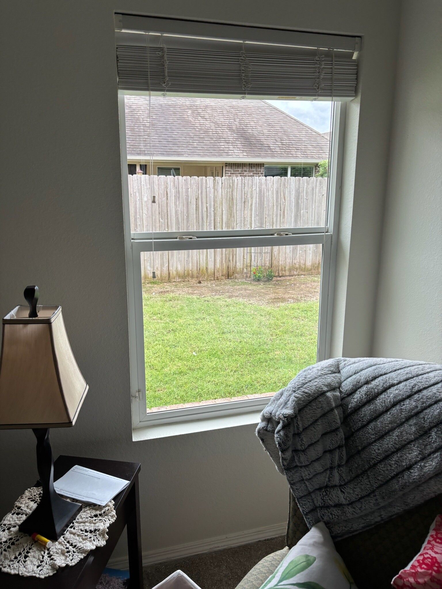 Window with open view of backyard with grass and a wooden fence. A lamp and armchair are nearby.