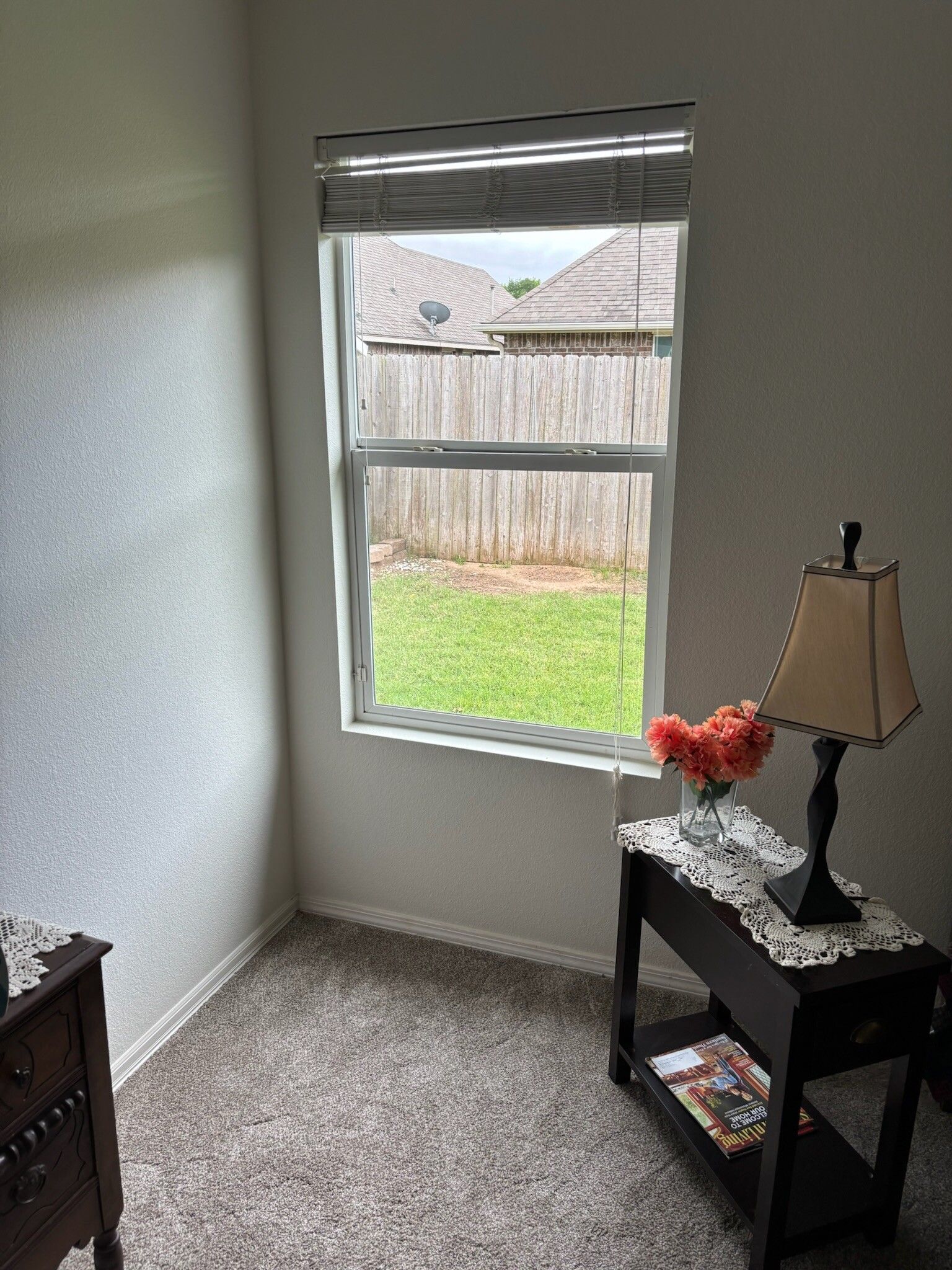 Small room with a window looking out at a yard. A table with a lamp and flowers sits below.