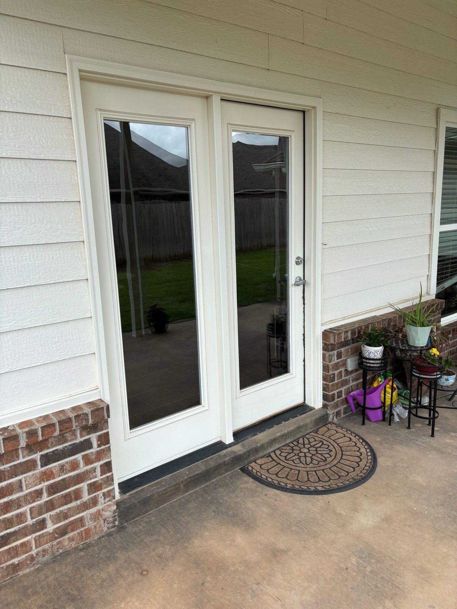 White double doors with glass panels on a porch, brick base, brown doormat, and plants.