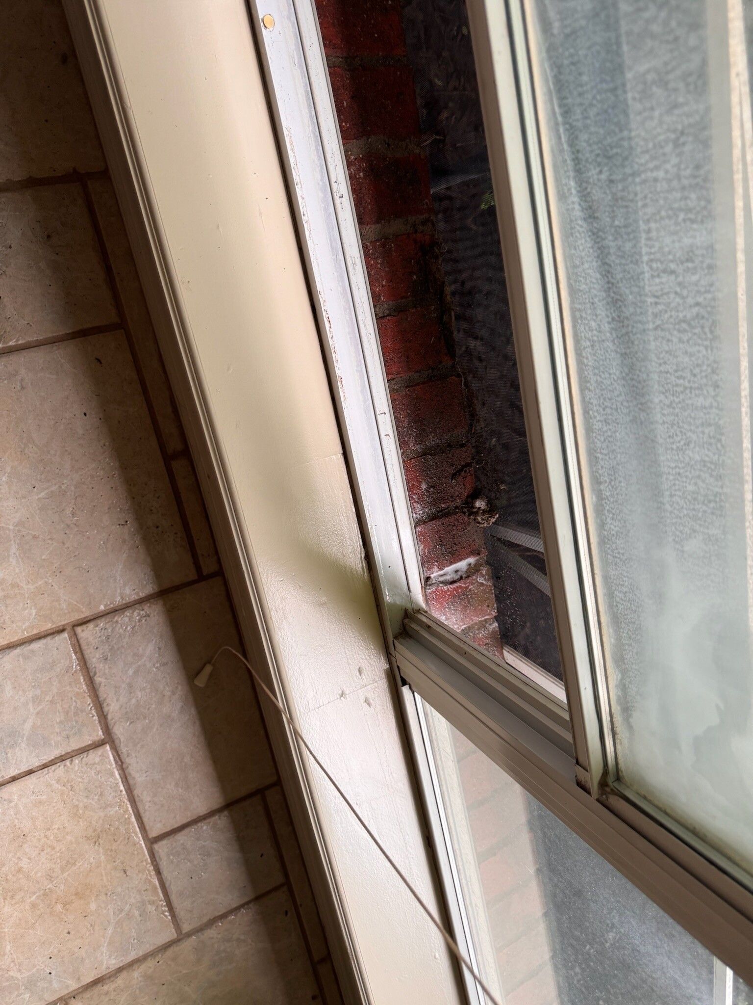 Close-up of a window frame with a dirty brick wall visible. The frame is beige and partially open.