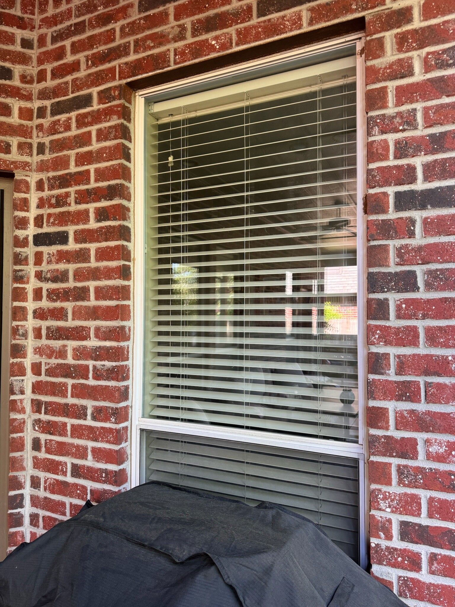 Window with closed blinds, set in a brick wall. A covered item sits below the window.