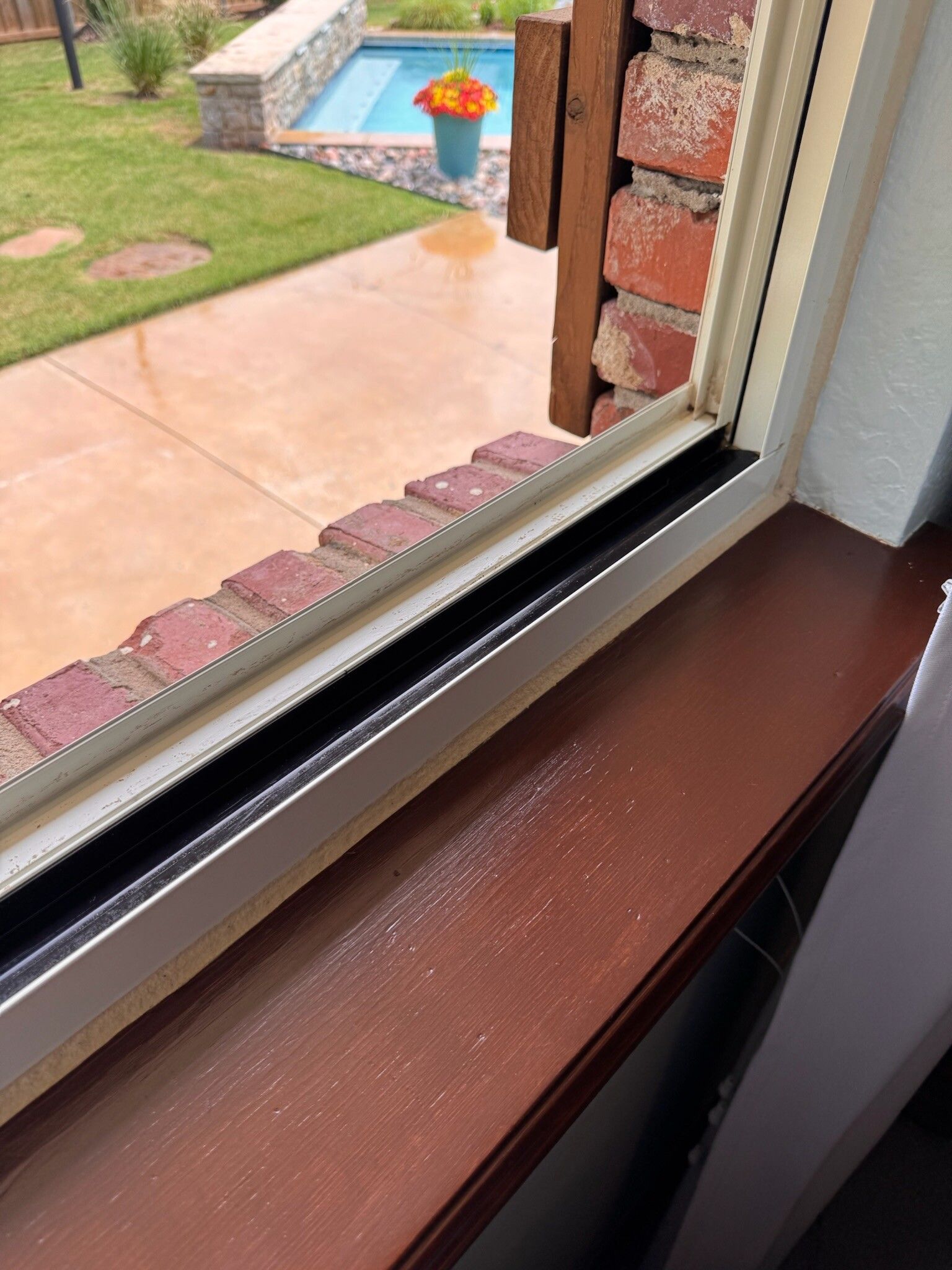 Wooden windowsill with view of a backyard, brick wall and small pool.