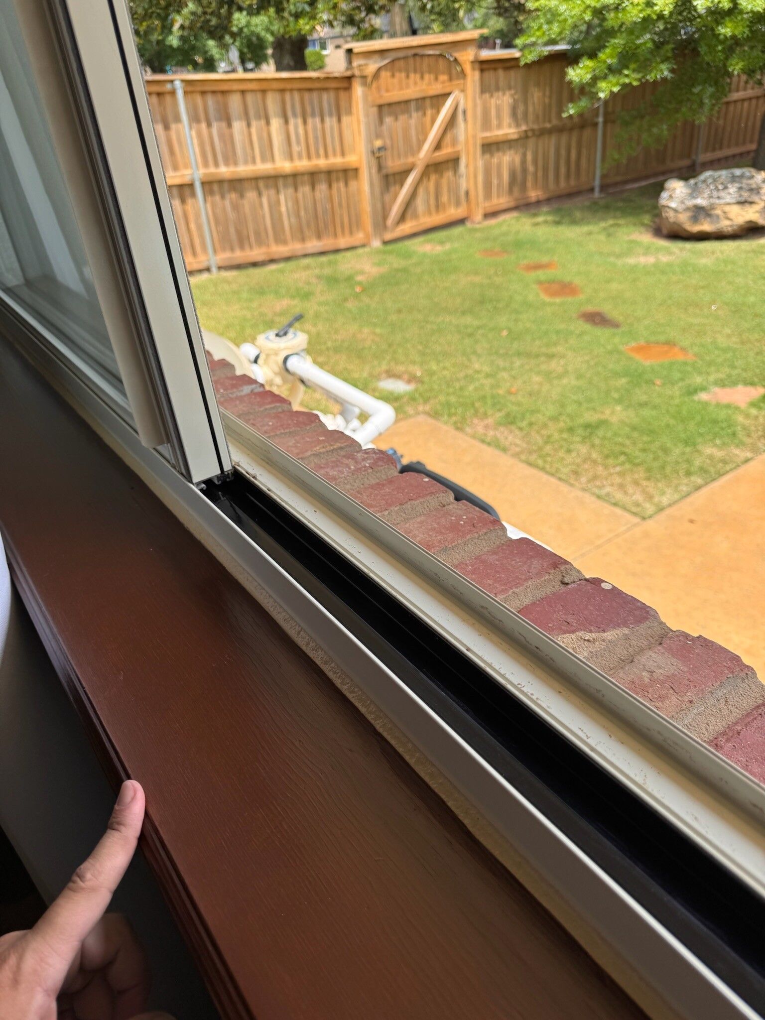 Finger pointing at a brown windowsill with a view of a backyard with green grass and a wooden fence.