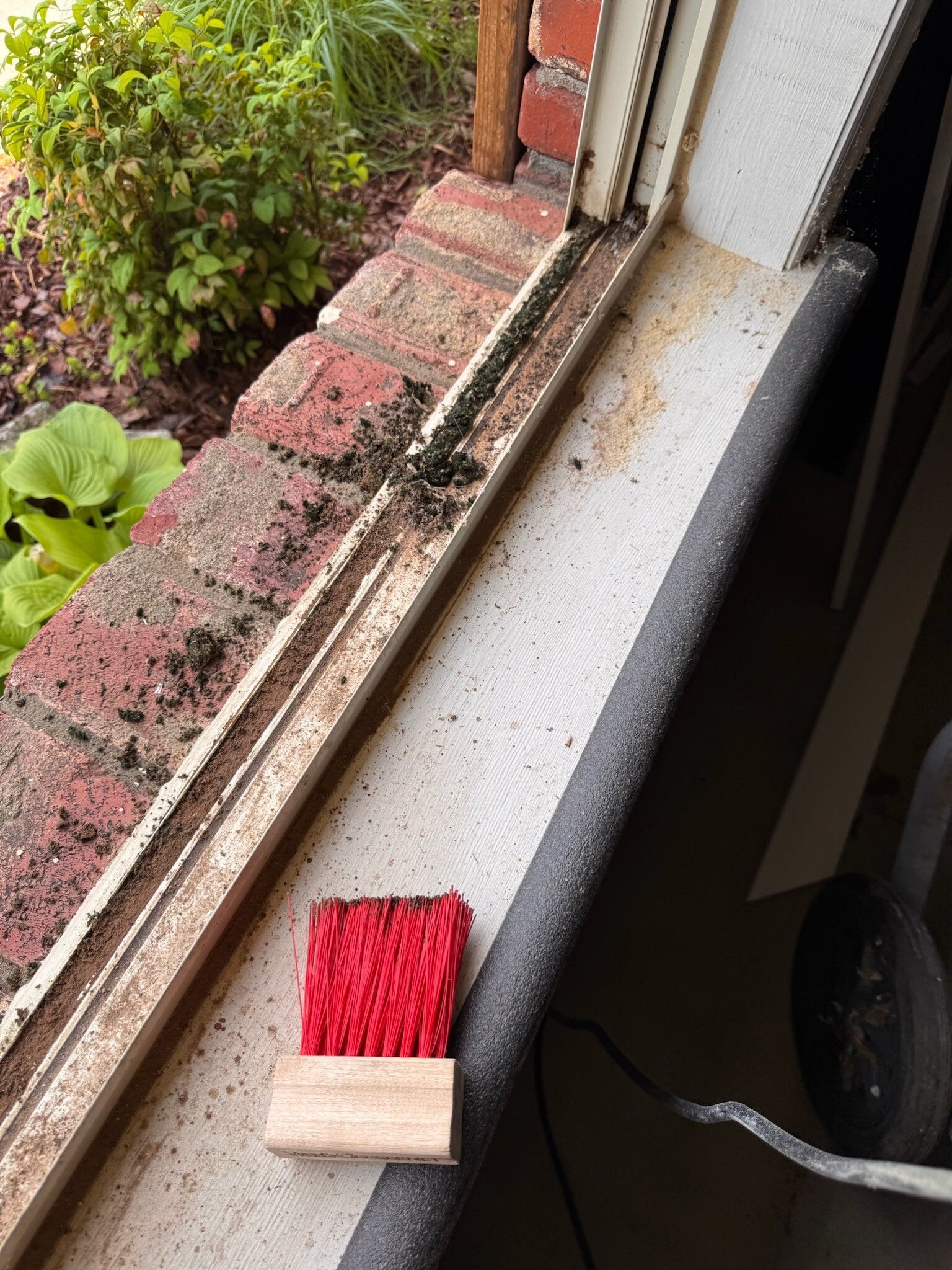 Window sill with a red scrub brush. Brick and white trim stained with dirt and debris.