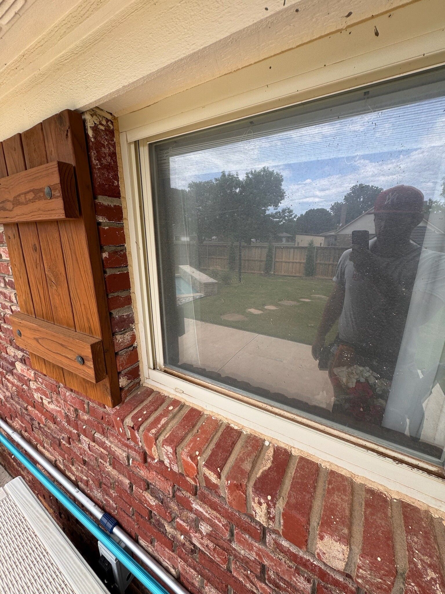 Window with screen; red brick exterior, wooden shutters, reflected figure holding a phone.