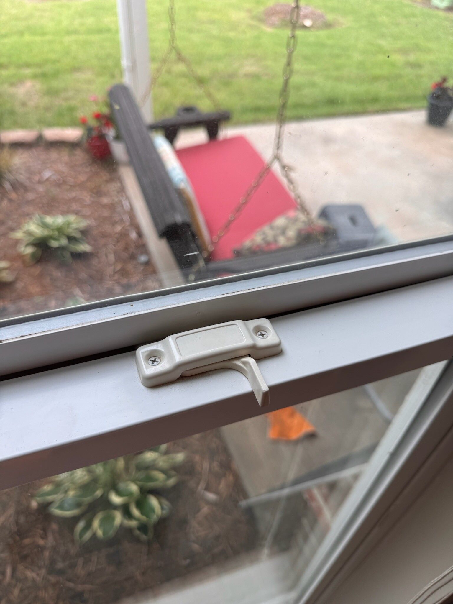 White window latch with a broken piece, set in a window frame with outdoor garden view.