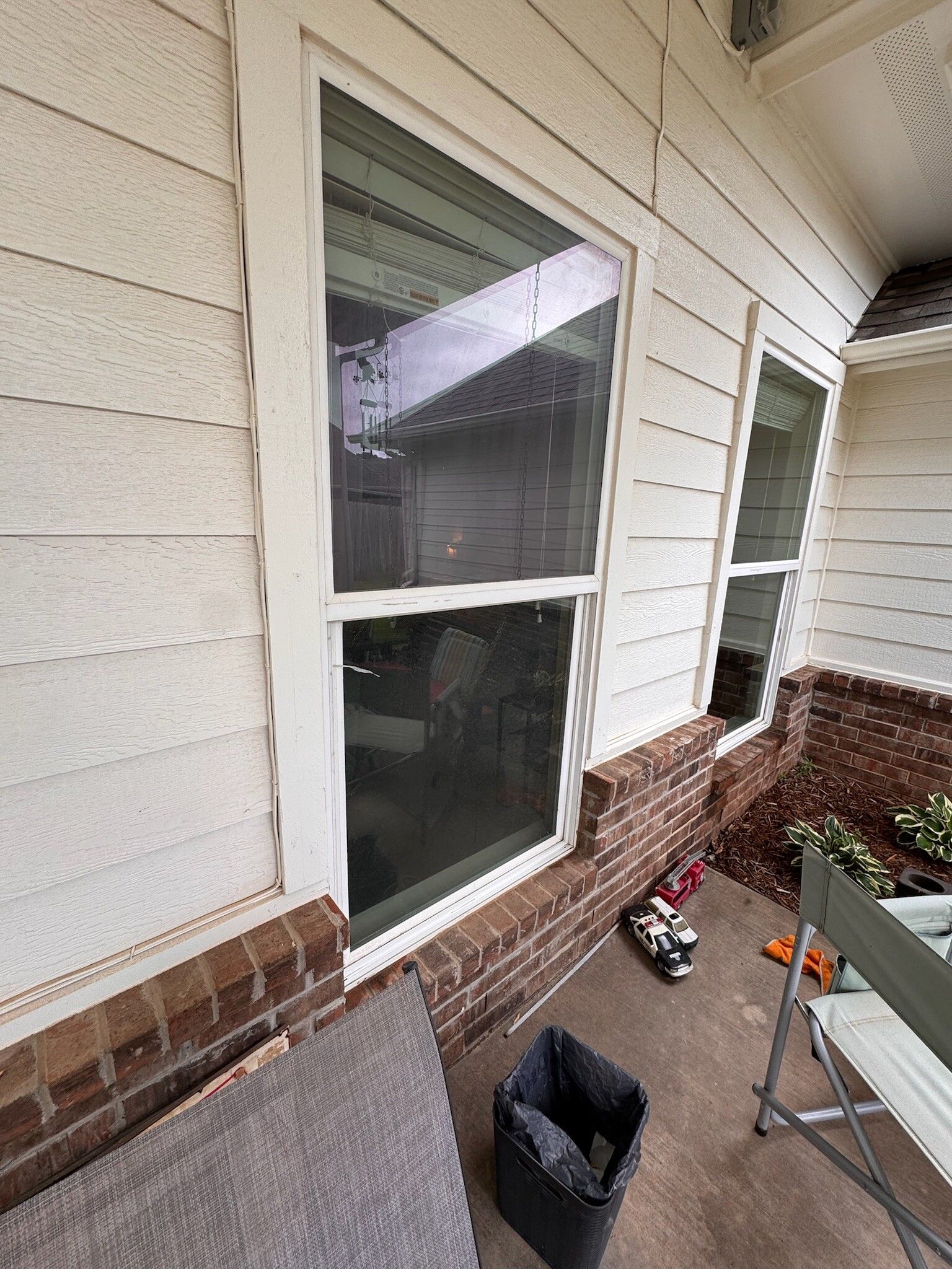 Two white-framed windows on a brick and siding exterior wall. A porch area with a chair and trash can is visible.