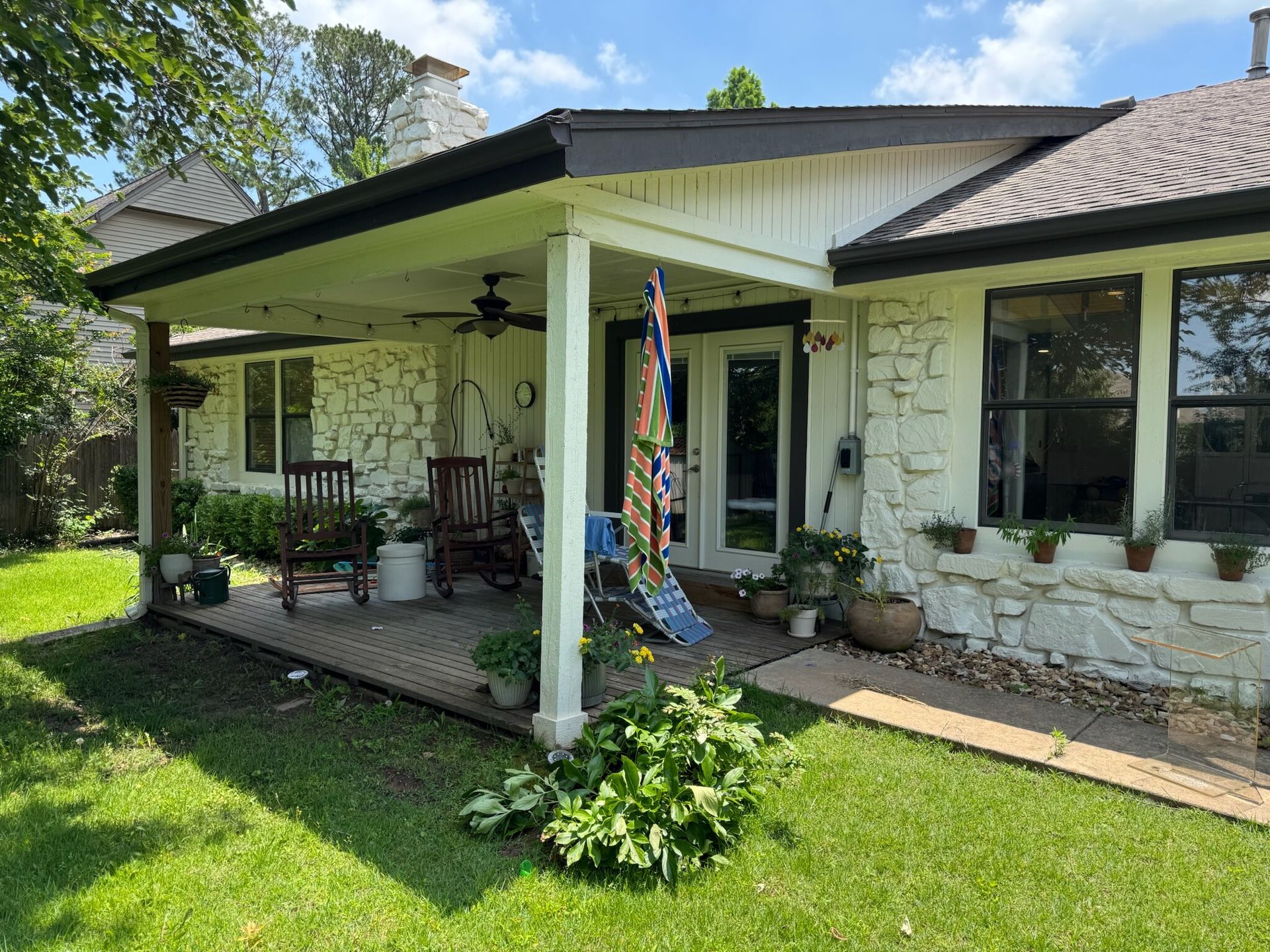 A house with a patio and lawn, featuring white stone walls, potted plants, and a rocking chair.