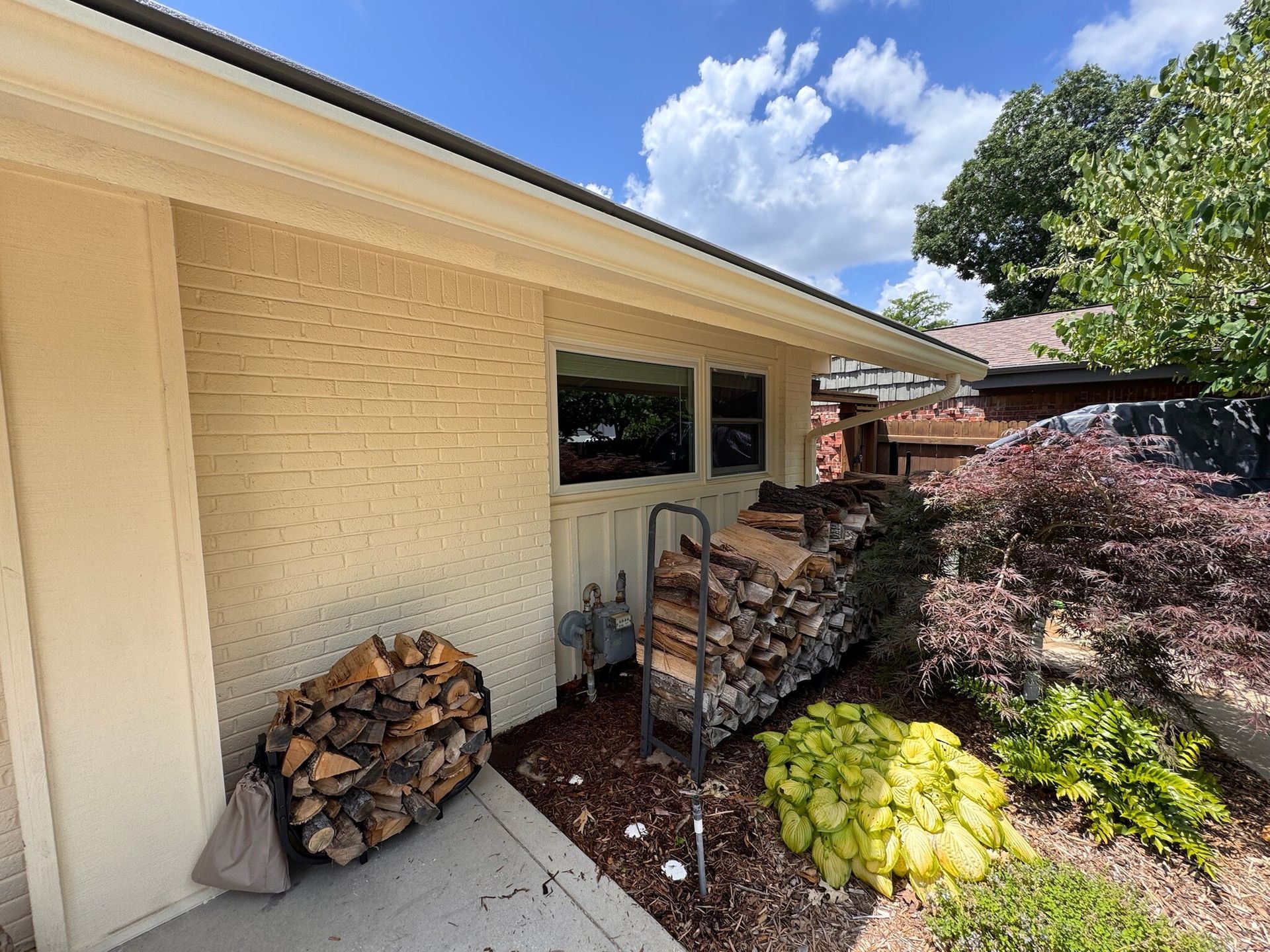 Exterior of a house with firewood stacked on a concrete path and garden. Brick wall, blue sky.
