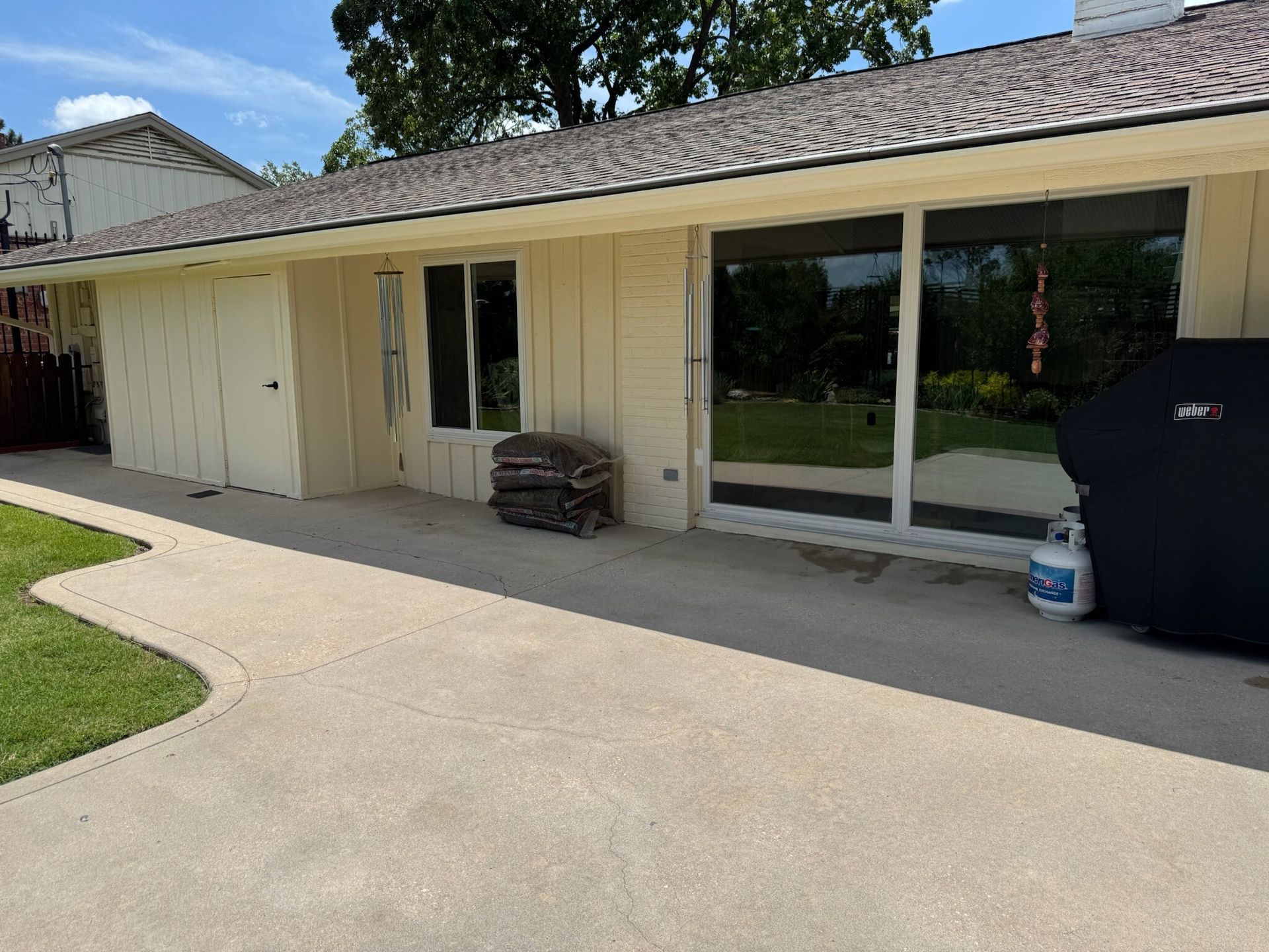 Beige building with a concrete patio. Large windows reflect a green yard. A grill and propane tank are visible.