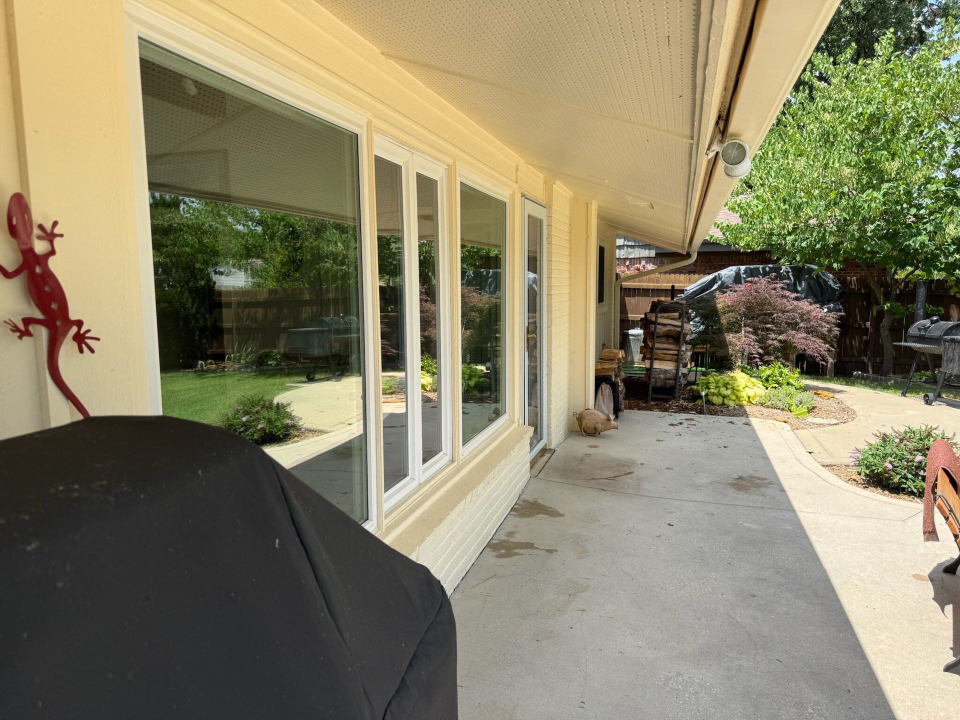 Patio with covered area, windows, grill, and decorative lizard on wall. Green foliage in background.