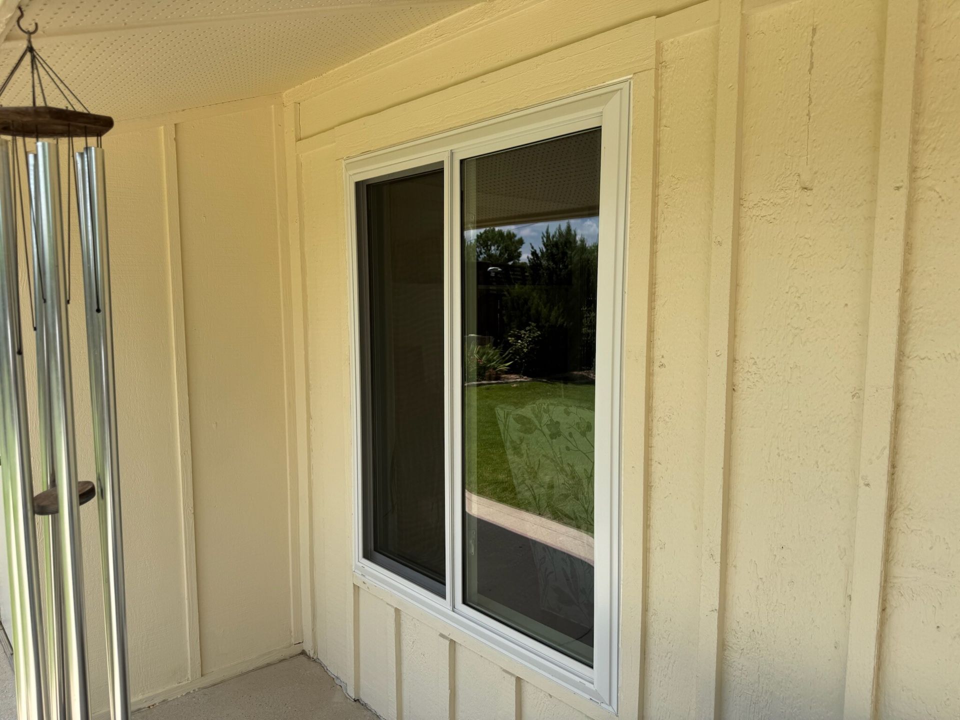 Window with white frame and dark screen reflects outdoor greenery, set in cream-colored siding.