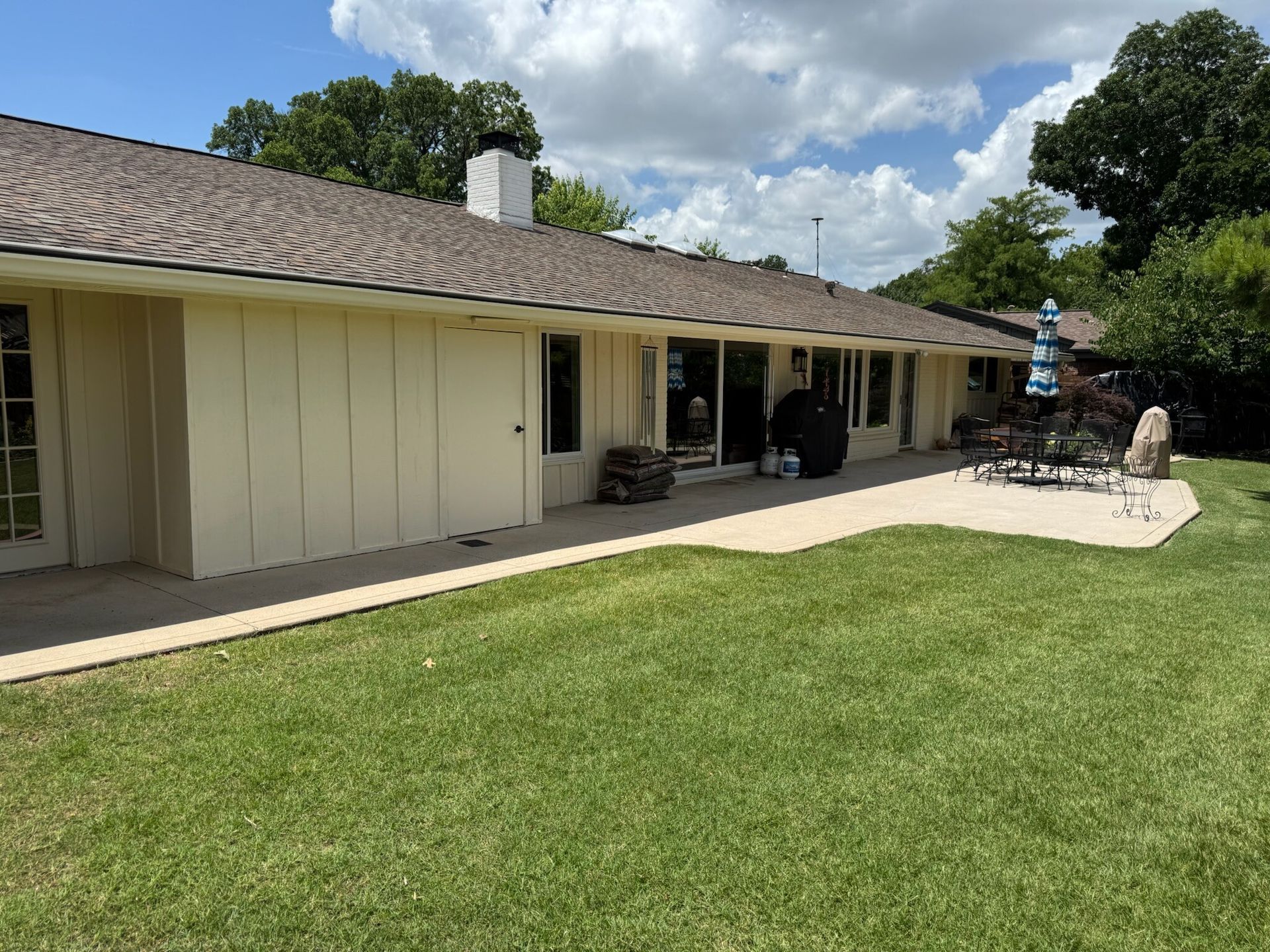 Back of a light yellow house with a concrete patio, green grass, and trees under a blue sky.