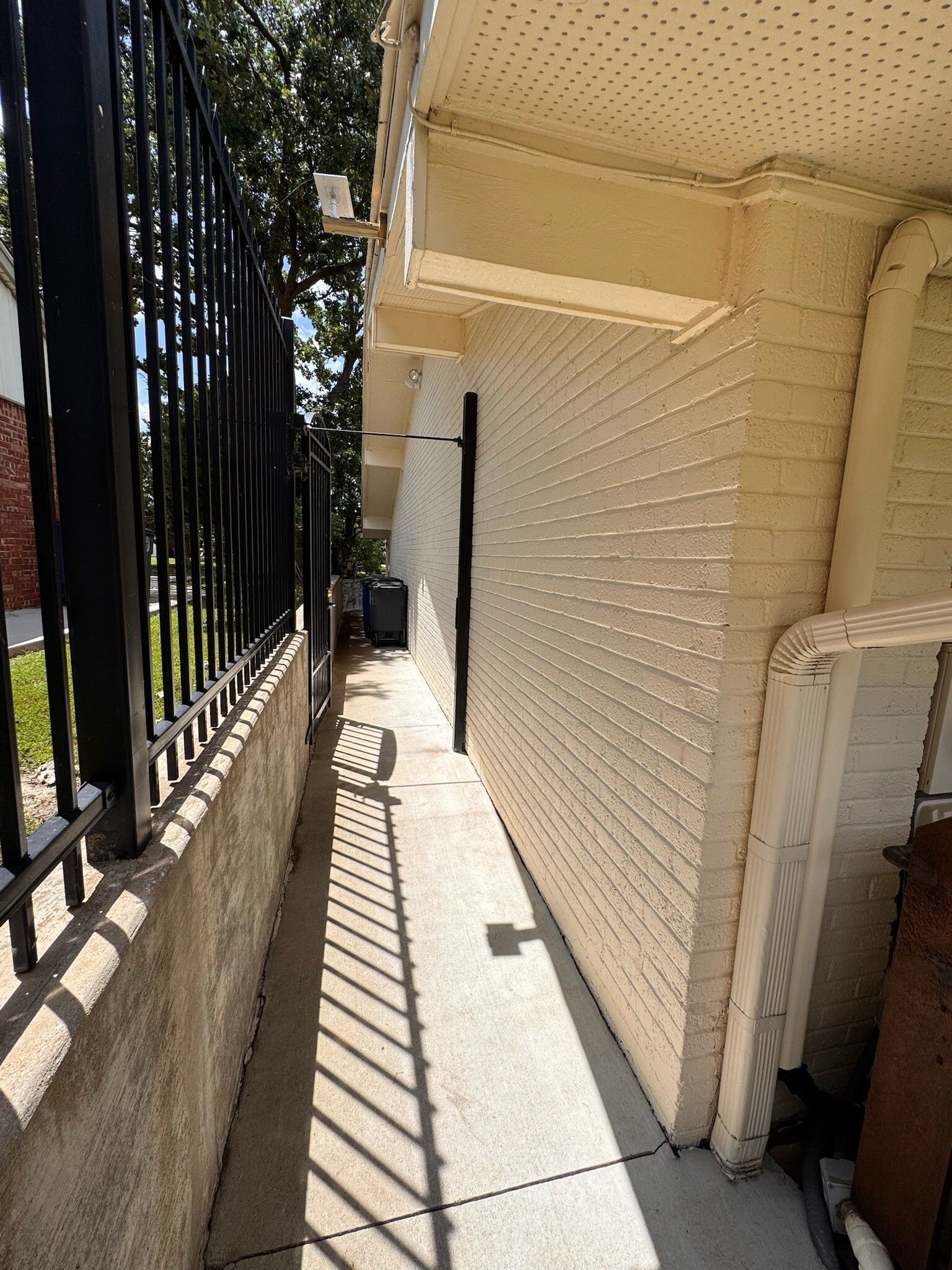 Narrow walkway between a black metal fence and a white brick building.