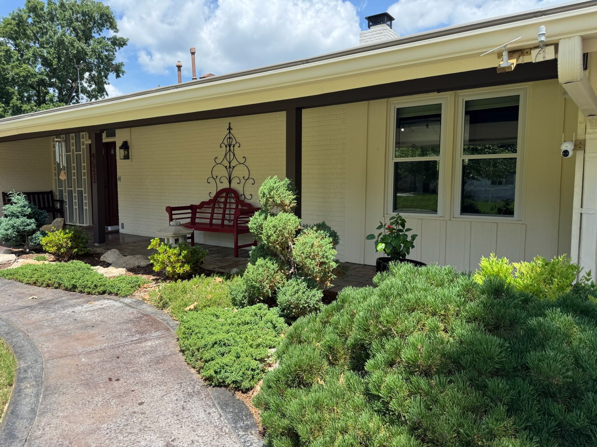 Front view of a house with a concrete walkway and landscaped garden. Green shrubs, a red bench, and windows are visible.