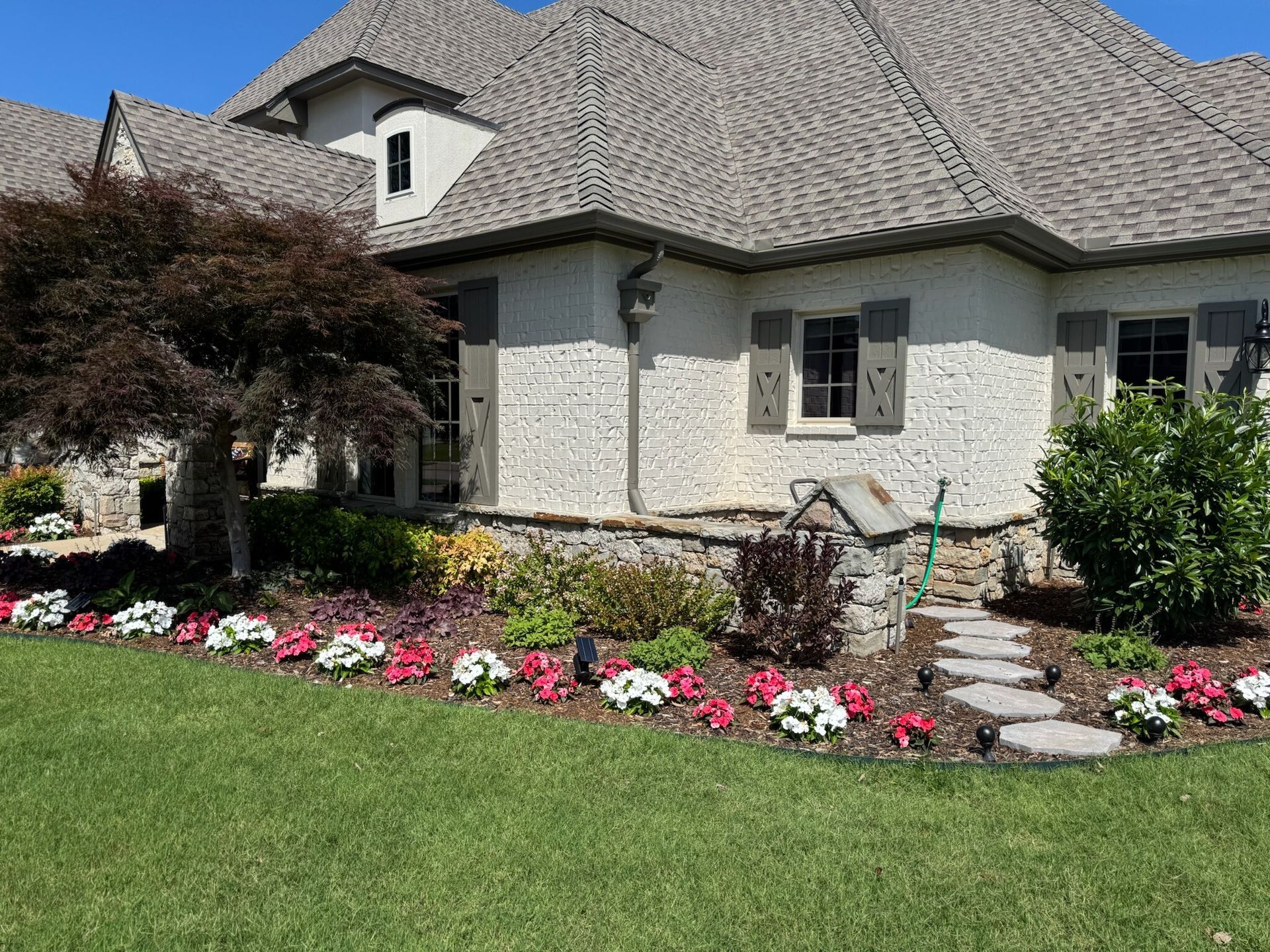 House exterior with flowerbeds and a walkway. Grey roof, stucco walls, and green lawn.