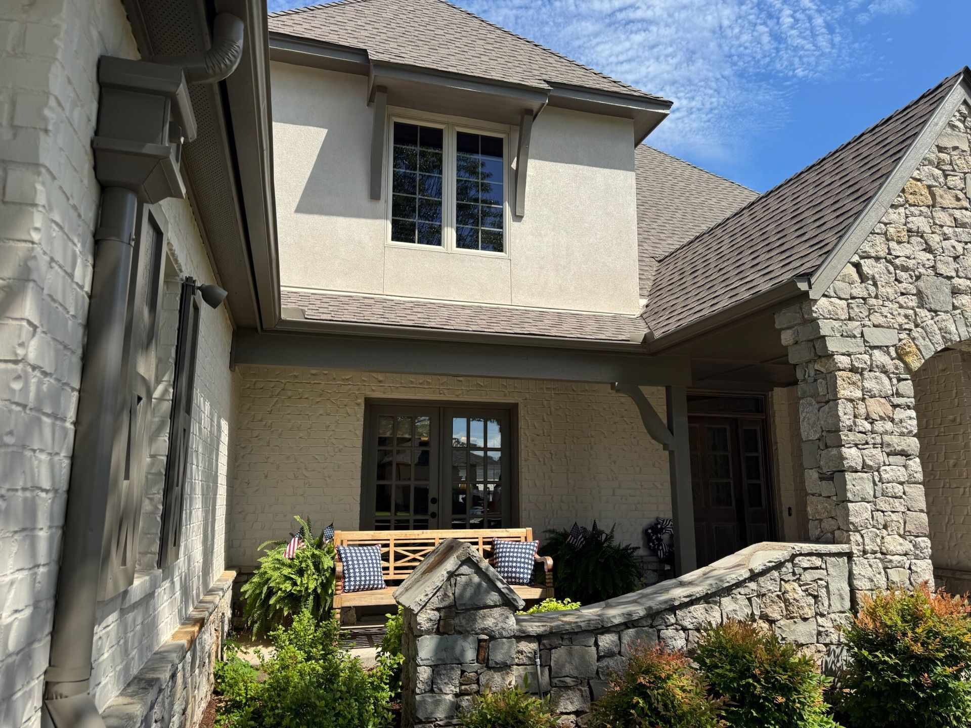 Stone and stucco house with dark-framed windows, porch, and a stone wall.