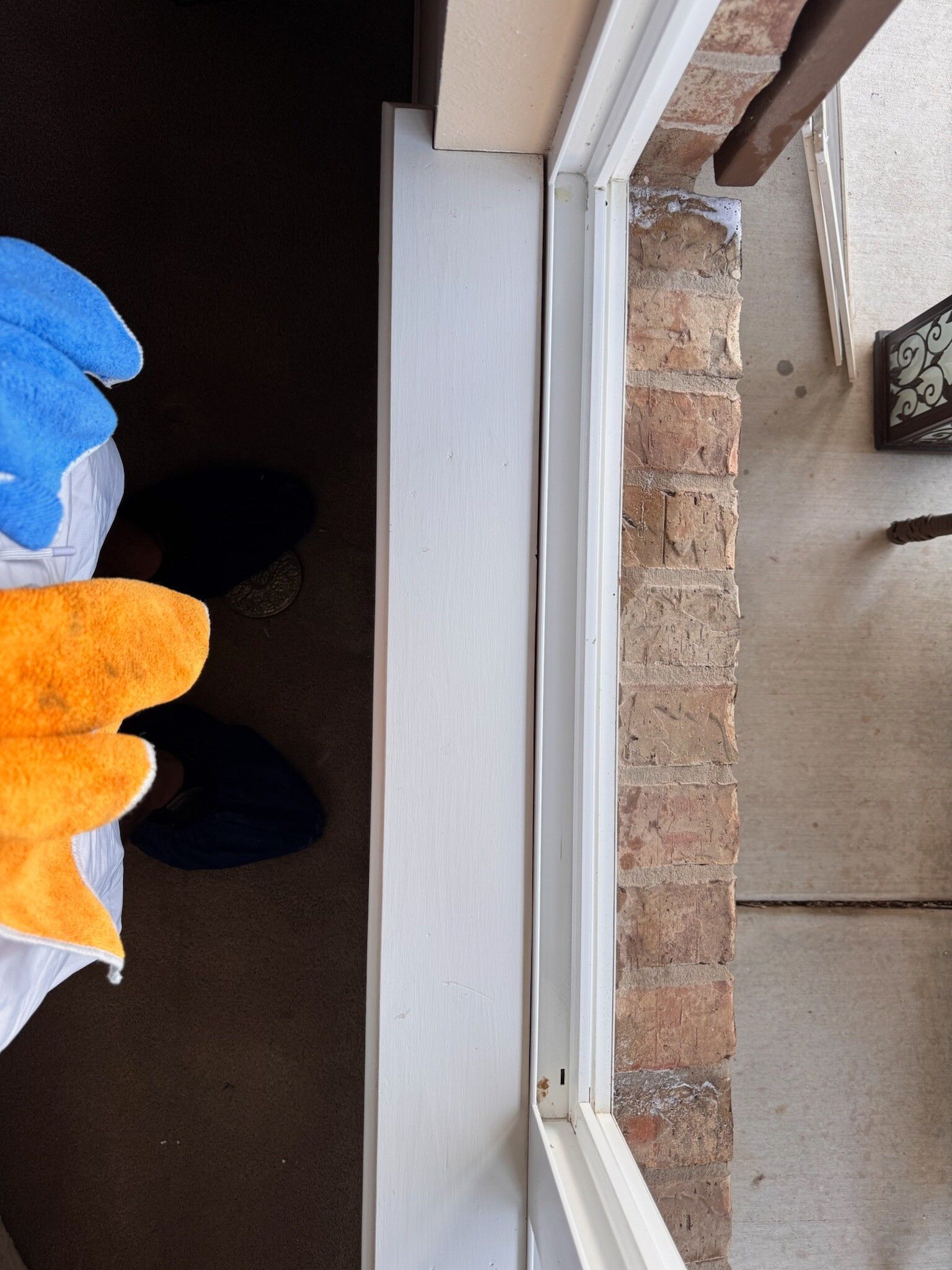 View of a white framed window against brick wall. Blue and orange gloves rest on a dark surface.