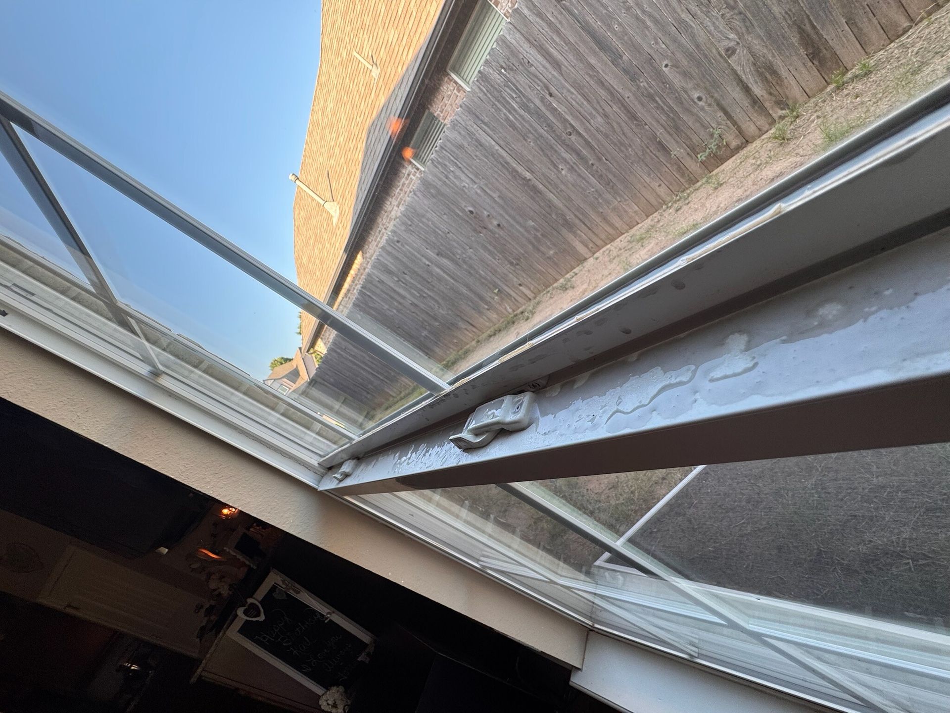 White-framed glass skylight, angled view from below. Light reflects off the glass.  Brick building and wood fence are visible.