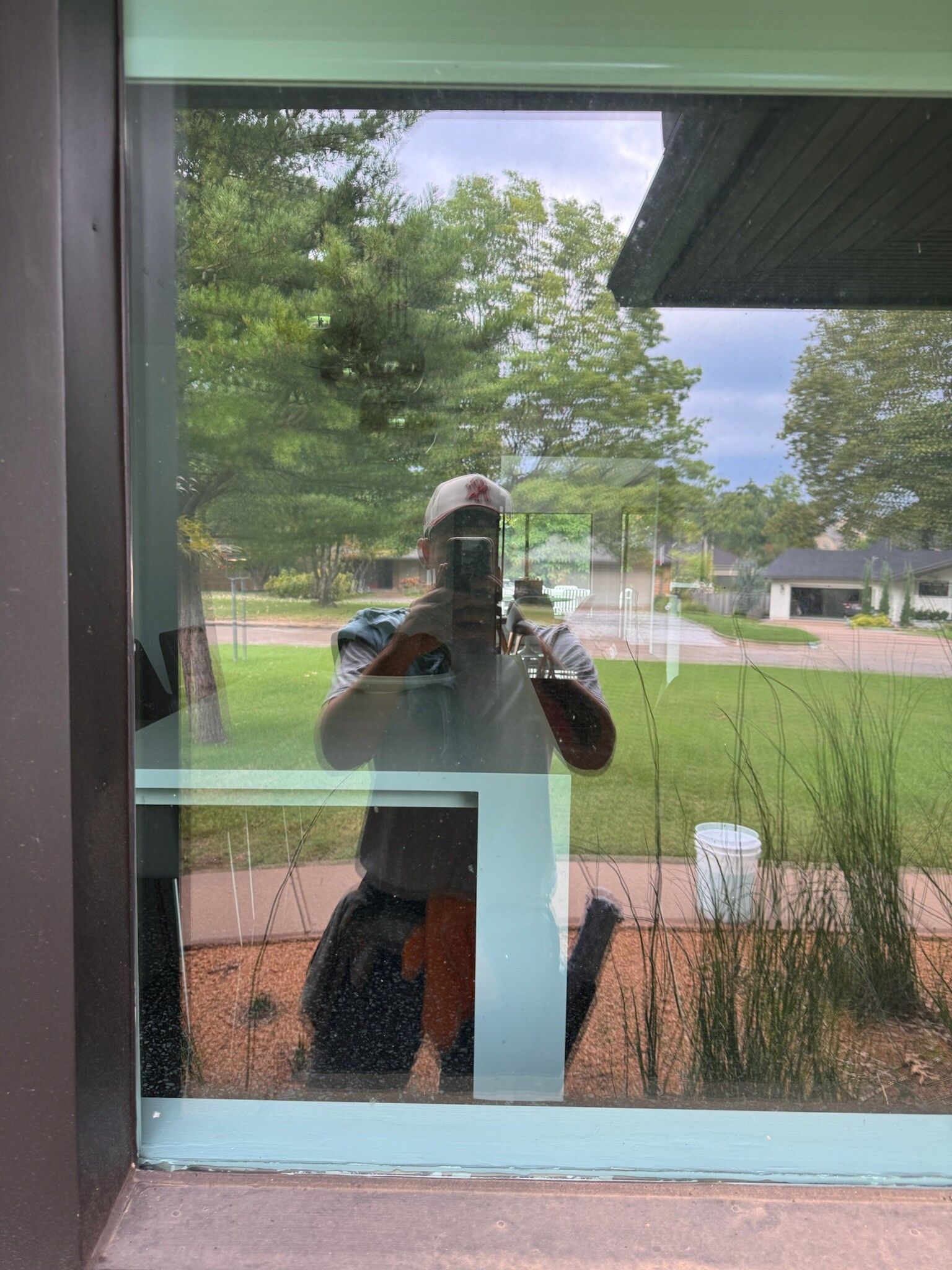 Person taking a photo, reflected in a window. Outside, trees, grass, and houses are visible.