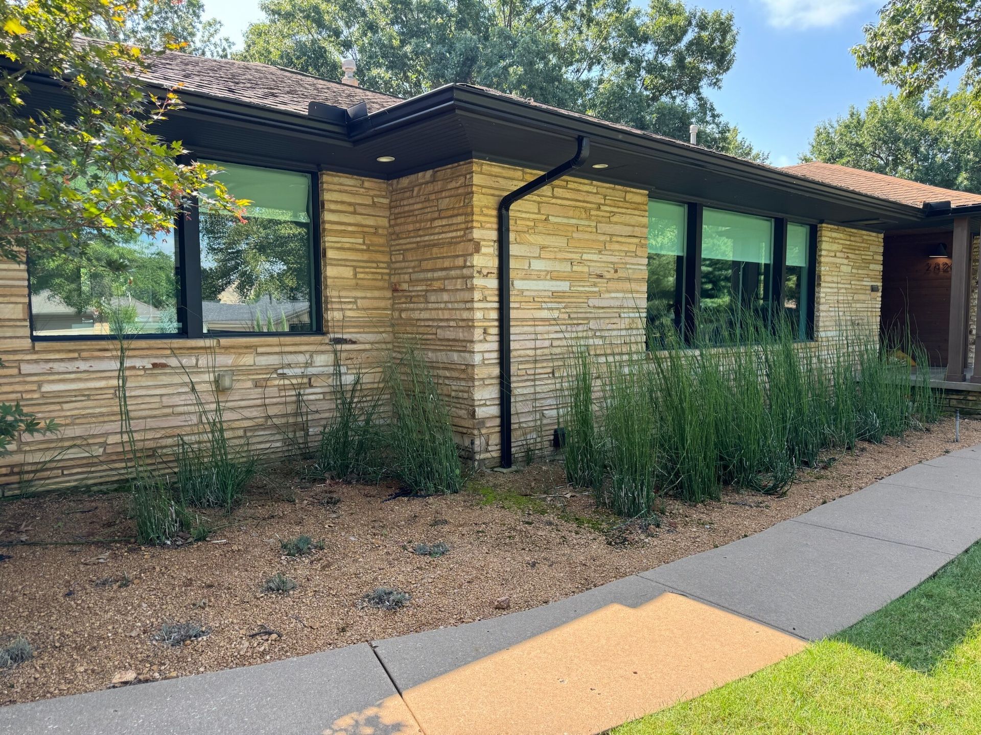 Tan brick home with black-framed windows, a dark roof, and a gravel walkway. Green plants line the foundation.