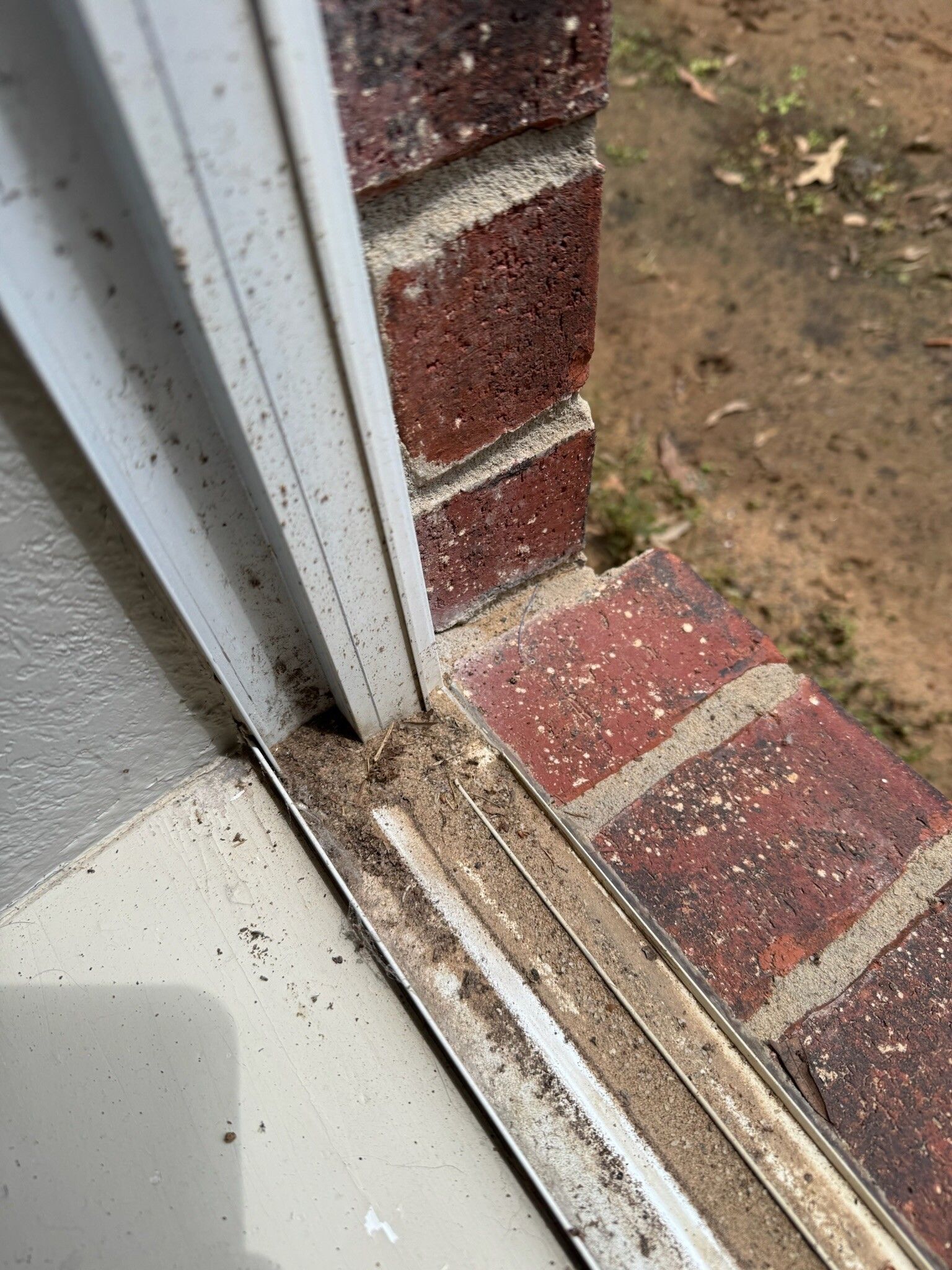Close-up of a window frame with debris accumulated in the bottom track, next to brick exterior.