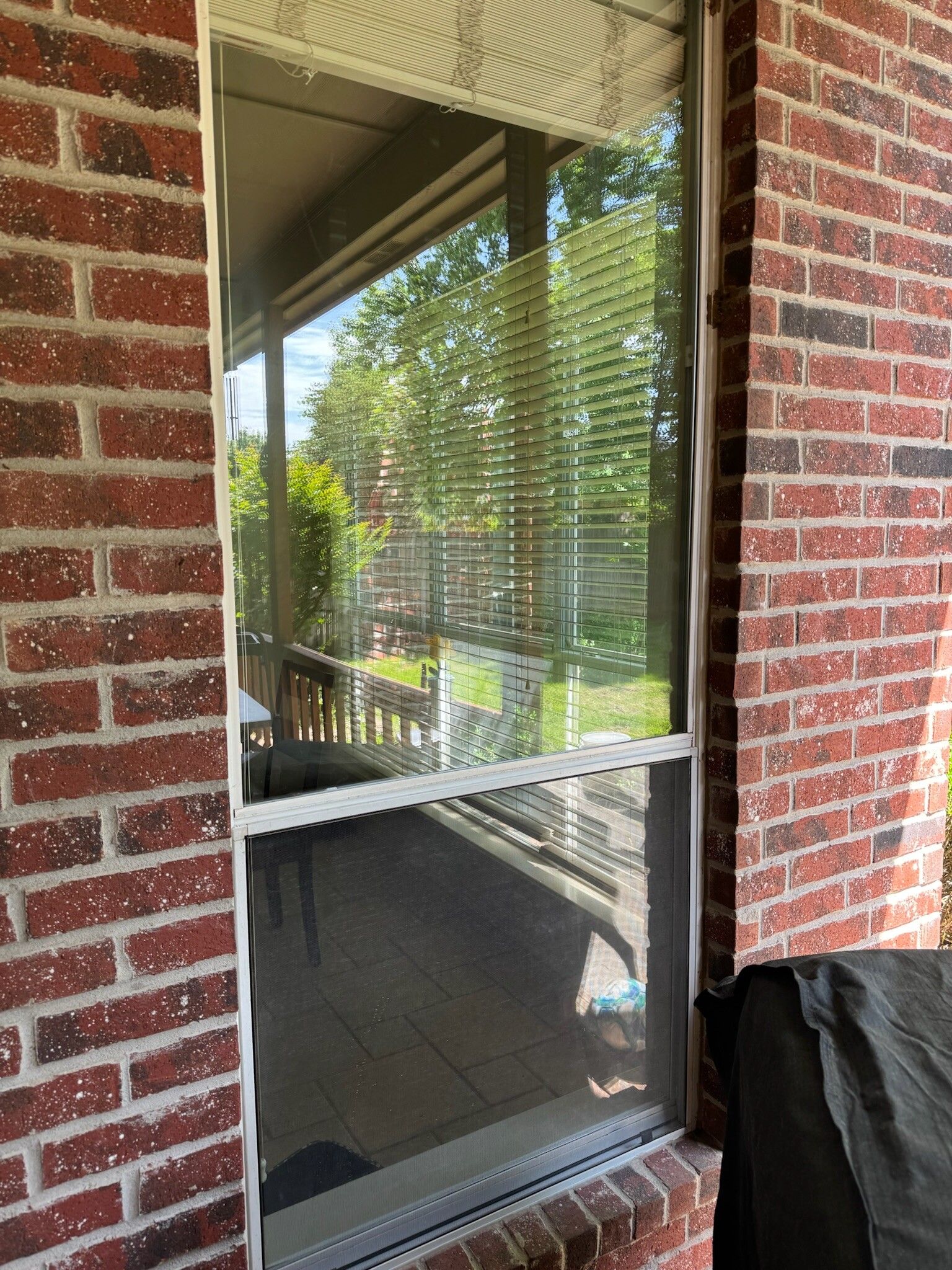 Window with screen and blinds, set in red brick wall. Interior view of deck with trees.