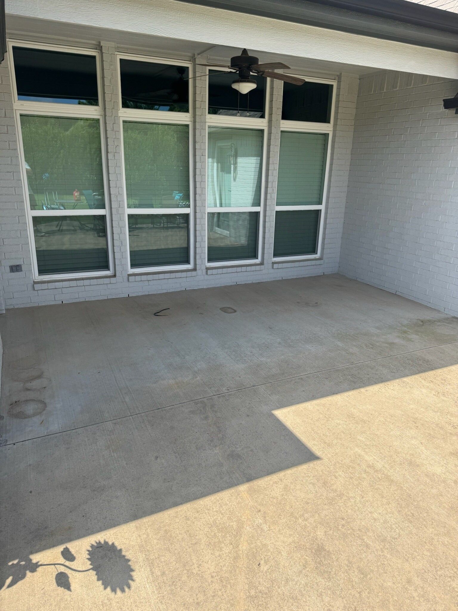 Covered patio with large windows and a ceiling fan, casting long shadows on the concrete floor.