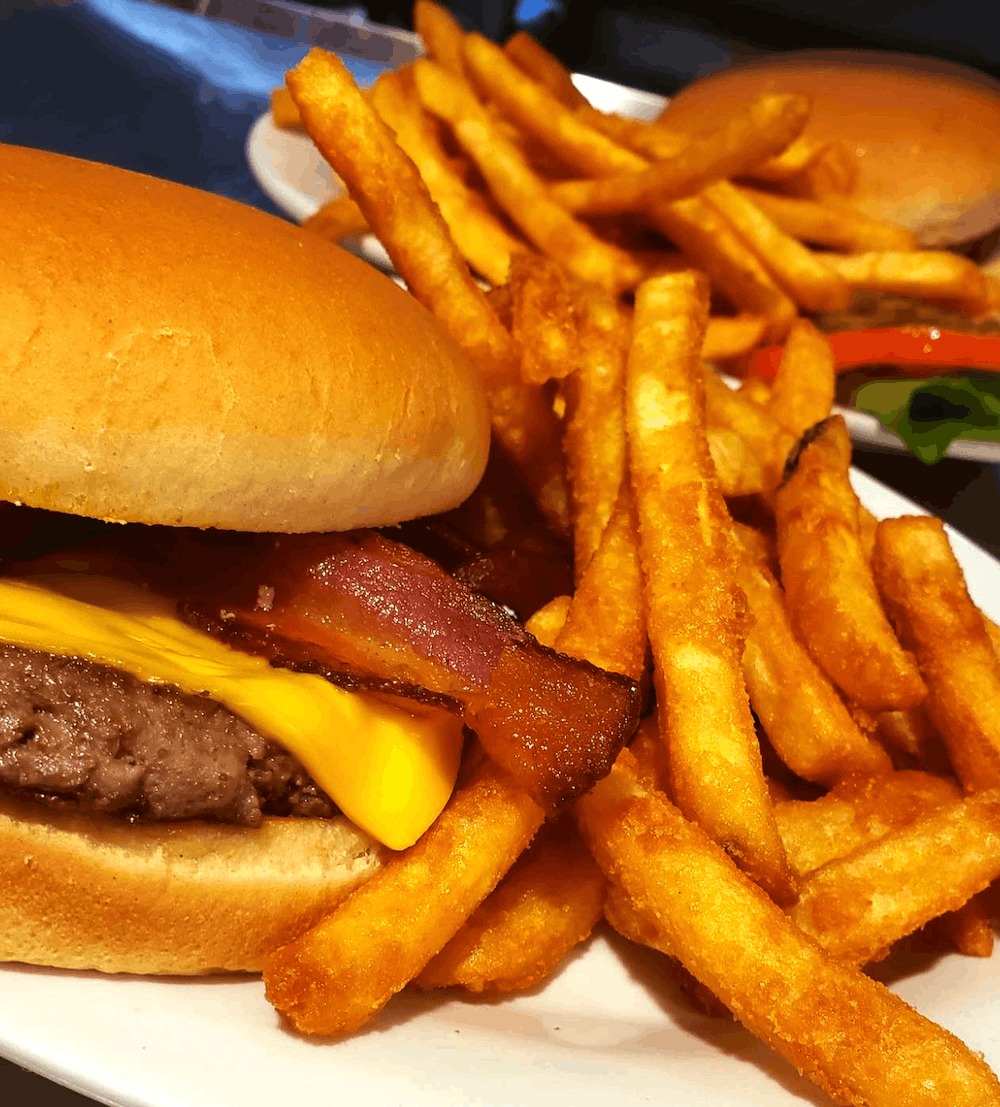 Cheeseburger with bacon and fries on a white plate. Second burger in background with lettuce and tomato.