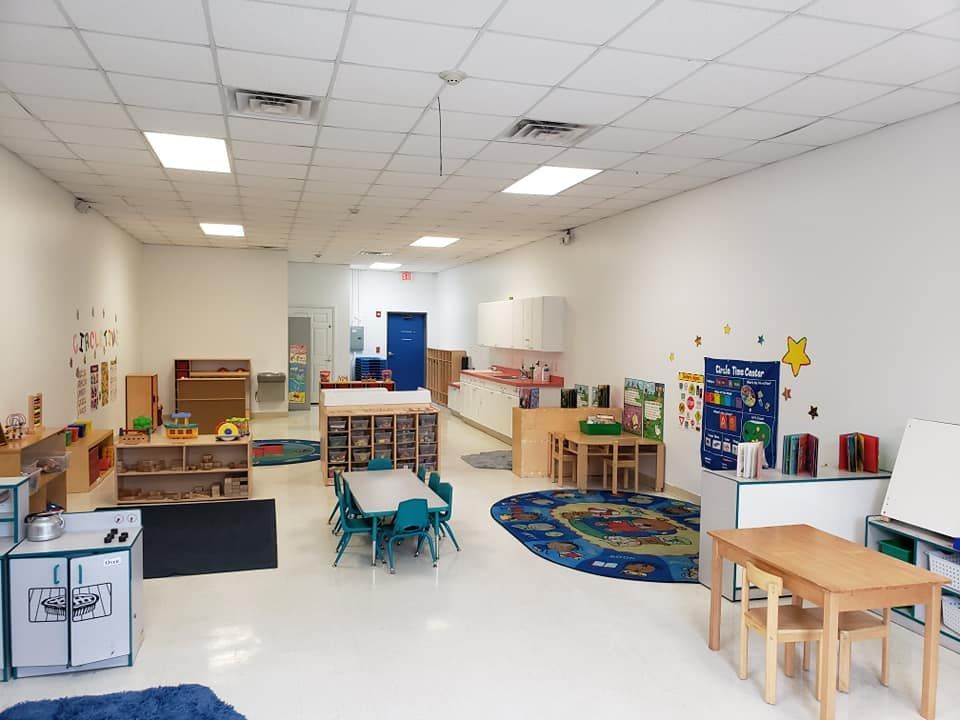 Empty, brightly lit preschool classroom with tables, toys, and shelving against white walls.