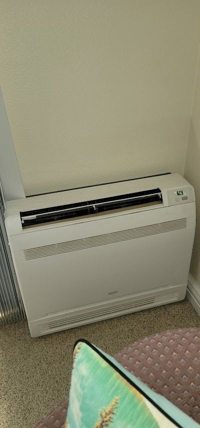 A white air conditioner is sitting on the floor in a living room next to a pillow.