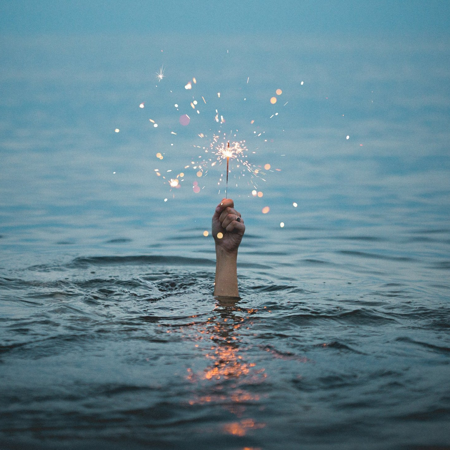 Hand reaching out of a body of water holding a sparkler up to keep it from getting extinguished.