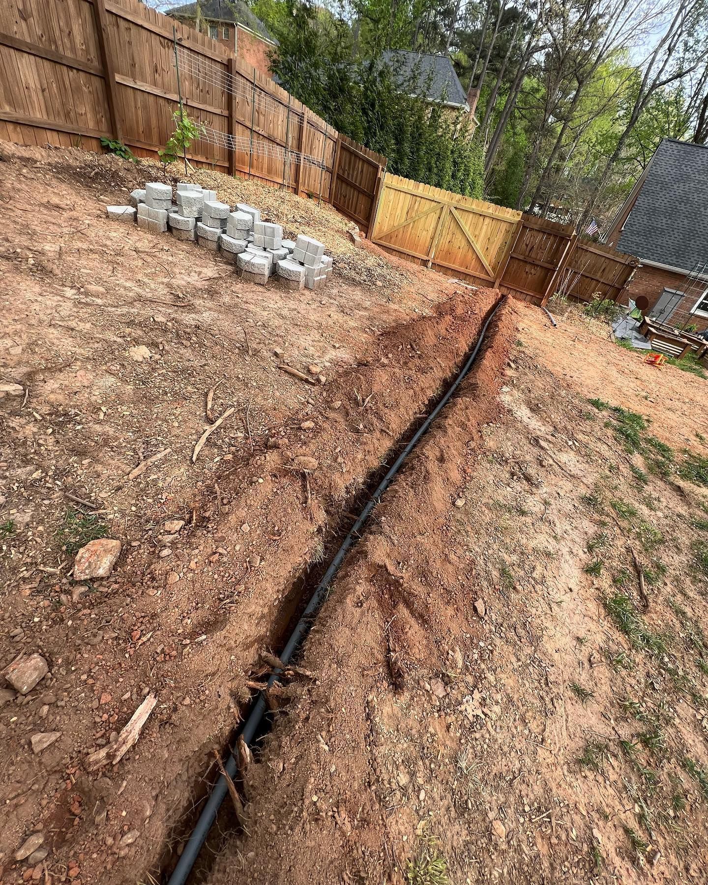 A black pipe is laying in the dirt in a backyard next to a wooden fence.
