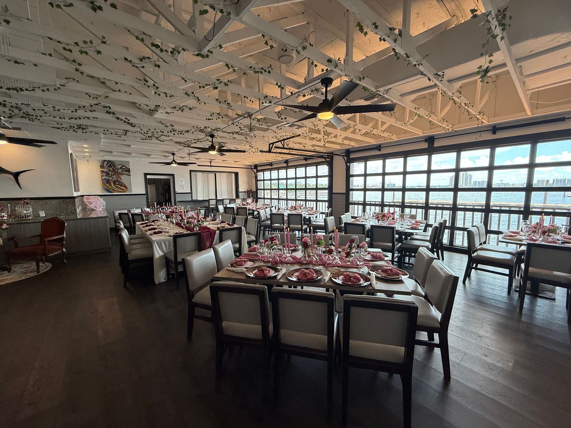 Dining room with long tables set for a meal, a high ceiling with vines, and windows overlooking a view.