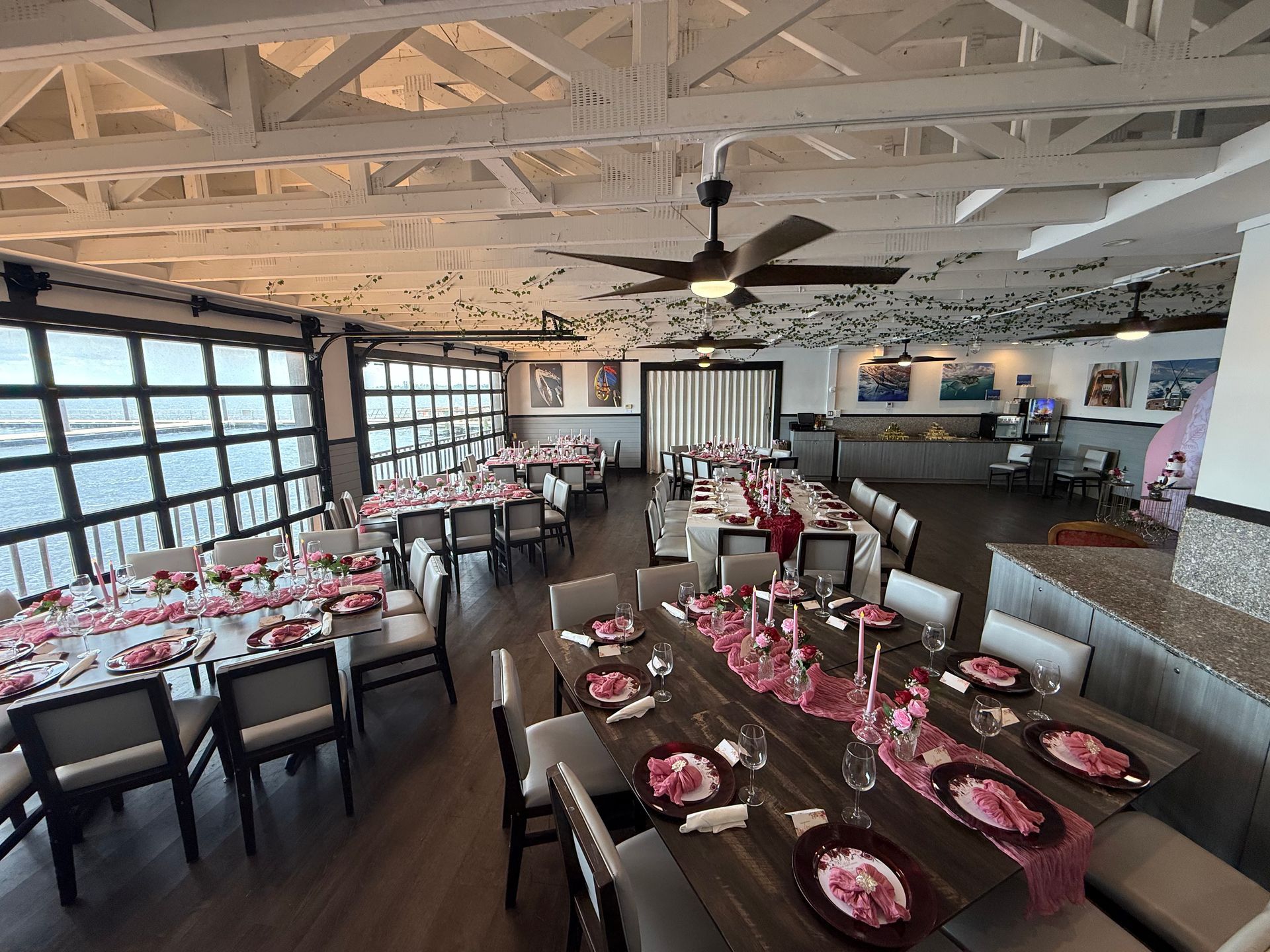 Dining room set for a celebration. Tables are decorated with pink runners, plates, and flowers. Large windows offer a view.