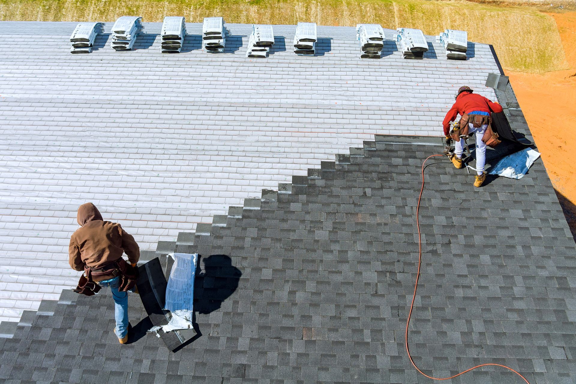 Roofing contractors install dark gray shingles on house roof using tools and bundled materials.