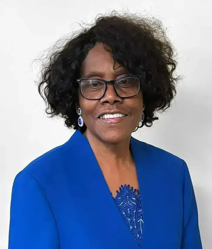 Woman with dark curly hair, glasses, and blue jacket smiling against a white background.