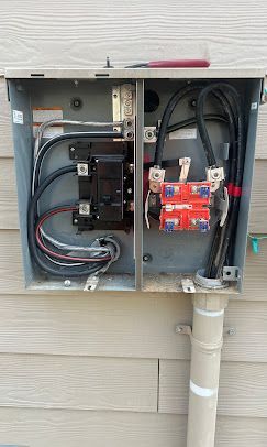 Electrical panel mounted on siding with wires and circuit breakers.
