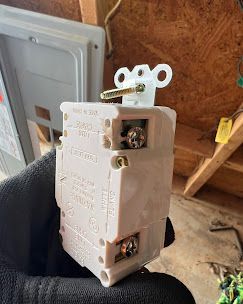 Close-up of a white electrical outlet held in a black gloved hand; visible screws, metal prongs, and wiring ports.