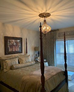 Bedroom with ornate four-poster bed, chandelier, painting, and window with blinds; soft lighting.