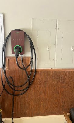 EV charger mounted on a beige wall with wooden paneling. Black cable coiled, green ring around plug.