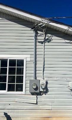 Exterior view of a light gray house with a window, power meters, and electrical wiring against a clear blue sky.