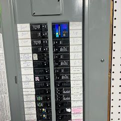Electrical breaker panel with circuit labels. Gray metal box, breakers mostly on, some labels handwritten.