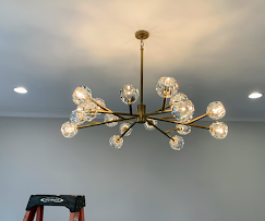 A modern, brass chandelier with crystal globe lights hangs from a white ceiling above a stepladder.