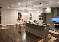 Kitchen under renovation: grey cabinets, island with tools, ladder, dark wood floor, pendant lights.