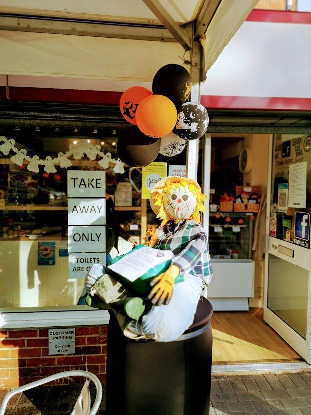 Scarecrow on a black trash can with balloons in front of a shop with a 