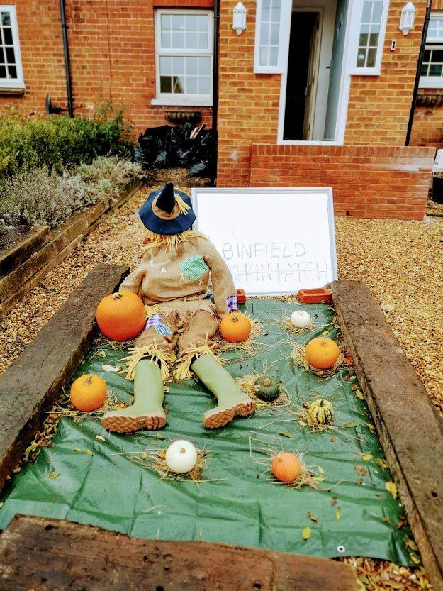 Scarecrow display in garden with pumpkins, straw, and a sign in front of a brick house.