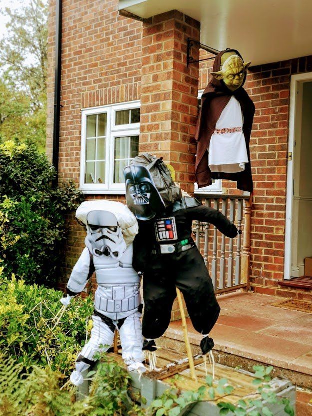 Star Wars-themed scarecrows on a porch: Yoda, Darth Vader, and a Stormtrooper. Brick house background.