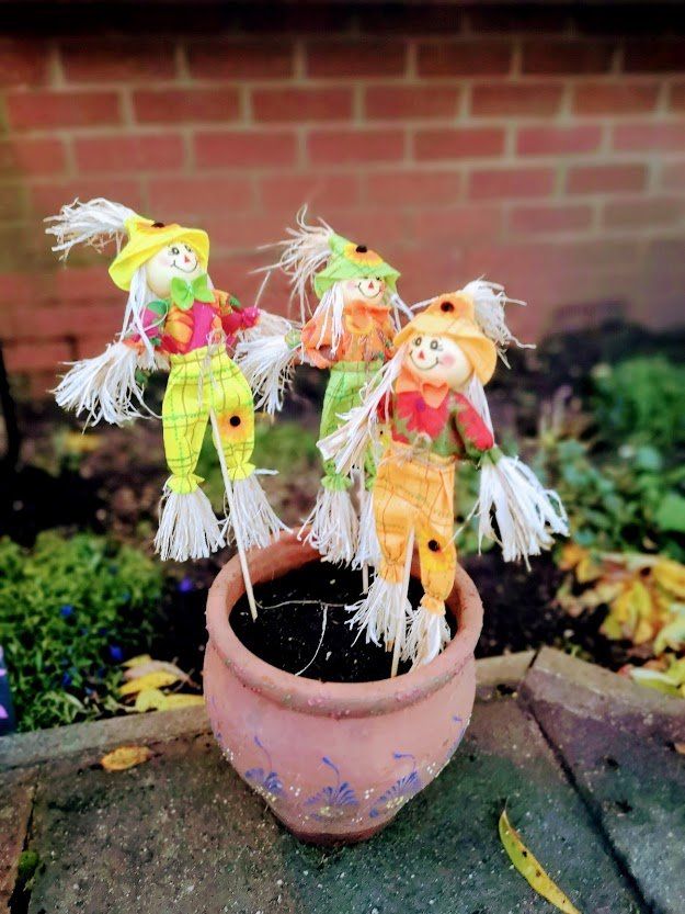 Three colorful scarecrow figures in a flowerpot against a brick wall.